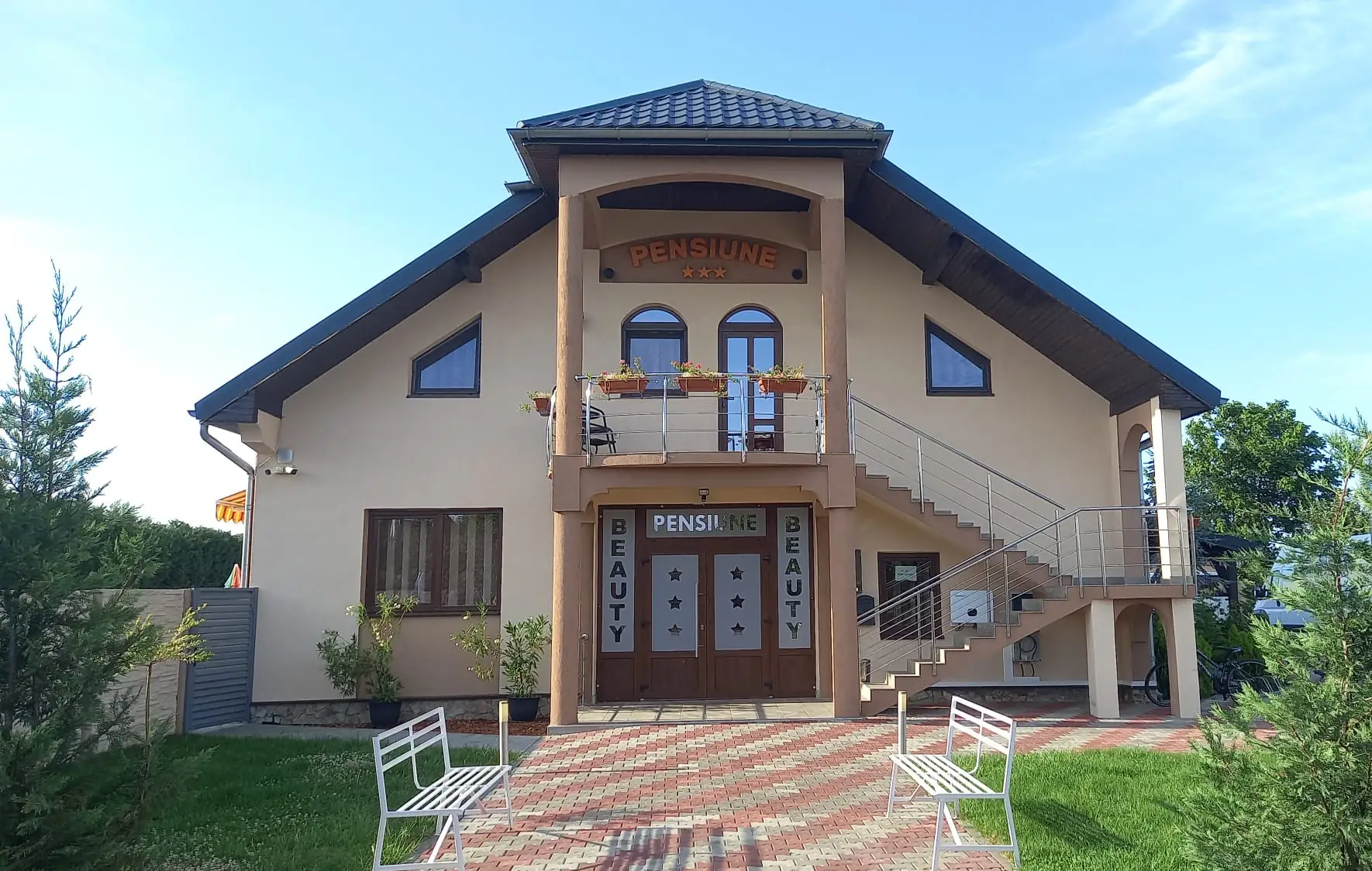 Fațada Pensiunii Beauty, cu firmă luminoasă, bănci albe și grădină, într-o dimineață senină