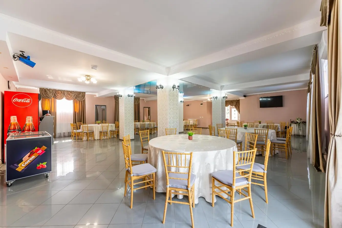 Sală de mese generoasă, cu mese rotunde, fețe de masă albe și scaune aurii tip Chiavari